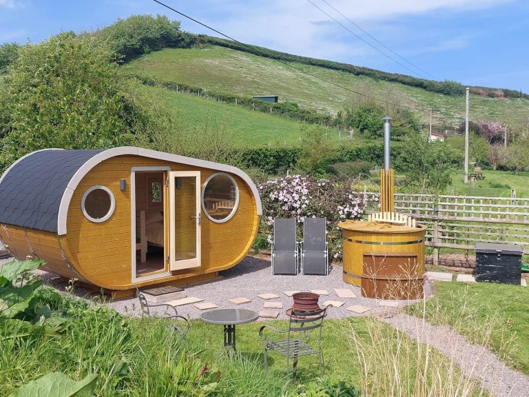 Modern glamping pod with circular windows, surrounded by greenery and a patio area.